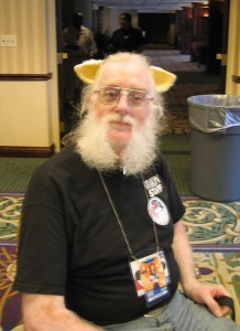 A man in a black T-shirt, wearing wolf ears, and with a full set of gray muttonchops, sits on a chair, looking at the camera